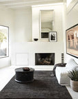 A modern, minimalist living room with white walls and fireplace, black circular coffee table, white sofa with gray blanket, Armadillo&Cos Savannah Rug - Carbon hand-knotted from a wool-cotton blend, framed art, and a potted plant in the corner.