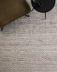 A textured beige Savannah Rug - Dust by Armadillo&Co with a subtle chevron pattern lies next to a dark olive chair and round black side table with a glass and bowl, all viewed from above. The hand-knotted wool cotton rug anchors the scene.