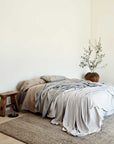 Minimalist bedroom with an unmade bed in neutral bedding, a rustic wooden stool, a glass of water on the floor, an Armadillo&Co Savannah Rug - Dust, and a potted plant in the corner against a plain white wall.