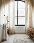 A bright bathroom features sheer beige curtains on an arched window, a white bathtub with draped towels, a woven round ottoman, and the Armadillo&Co Savannah Rug in Dust for added floor texture.