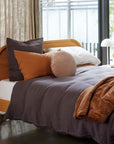 A modern bed with a wooden headboard features gray linens, an orange-brown blanket, and CITTA’s Velvet Round Cushion in Scoria Tint among other soft pillows, positioned next to a window and a small white lamp on the bedside table.