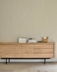 The Ethnicraft Shadow Sideboard with Legs - 71 stands against a plain beige wall, topped with neatly arranged magazines, books, a brown paper bag, and glasses.