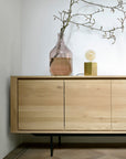 The Ethnicraft Shadow Sideboard with Legs - 88, crafted in European oak, stands against a white wall with a glass vase holding a bare branch and a small brass-based table lamp with an exposed bulb placed on top.