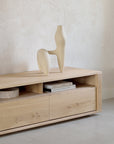 The Ethnicraft Shadow TV Cupboard - 55, a minimalist console in European oak, features two drawers and an open shelf for books, topped with a modern abstract beige sculpture against a light textured wall.