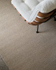 A close-up of a cream-upholstered chair with wood accents and black legs sits on the Sherpa Weave Rug - Chai by Armadillo&Co, beside a polished concrete floor.