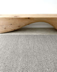 A modern wooden bench with a wave-like arch sits on the Sherpa Weave Rug - Pumice by Armadillo&Co, featuring a gray textured pattern against a white wall.