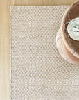 A beige textured woven placemat on a light wooden table, accented by a round bowl with an air plant—perfectly complementing the Sherpa Weave Rug - Sand from Armadillo&Co.