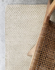 A close-up of the Armadillo&Co Sherpa Weave Rug in Sand on a concrete floor, with a wooden chair featuring a woven seat visible in the upper right corner.