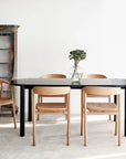 A modern dining room with a Cove Dining Table - Black Oak by Wooden Stories, four light wooden chairs, a glass vase of greenery, and an antique glass-front cabinet. Natural light highlights the inviting, minimalist atmosphere.