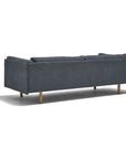 A dark gray Sonder Sofa | Fabric by Wooden Stories, featuring a modern linear frame and wooden legs, is shown from the back at an angle. The sofa highlights clean Danish-inspired lines and three feather-filled back cushions, casting a shadow on the white background.