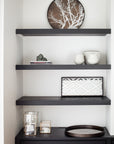 A minimalist black shelving unit with three shelves displays decorative items from the Ethnicraft Tray Collection, including the Slice Mirror Tray Light Aged - Round / Small, white vases, books, a geometric frame, and glass boxes with candles.