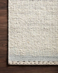 A close-up of the Loloi Sloane Rug - Mist showcases its hand-woven, light tone and stitched border on a dark wooden floor, highlighting subtle color variations and a simple, modern bohemian design.