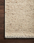 A close-up of the Sloane Rug - Natural by Loloi shows its textured, off-white hand-woven design with subtle flecks, set on a dark wooden floor for a modern bohemian vibe.