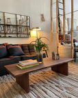 A cozy living room with a dark sofa and the Sofia Live Edge Coffee Table - Walnut by SOBU holding books, a plant, and decor on a textured rug. Wall art, a lamp, mirror, and wooden shelves complete this modern, inviting space.