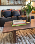 A cozy living room features the SOBU Sofia Live Edge Coffee Table in walnut, topped with a potted plant and art books, set before a dark gray sofa with patterned cushions on a textured rug. A large mirror shows additional chairs in the background.