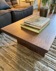 The Sofia Live Edge Coffee Table - Walnut by SOBU, crafted in solid American walnut with a satin lacquer finish, stands on a textured rug with three books. A gray upholstered couch and green potted plant complete the sunlit scene.
