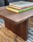 A close-up of the SOBU Sofia Live Edge Coffee Table in American walnut with a satin lacquer finish, three books stacked on top. The table rests on a textured beige rug, with part of a dark couch visible in the background.