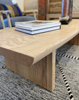A close-up of the Sofia Live Edge Coffee Table - White Ash by SOBU, crafted from solid American ash with a satin lacquer, stacked books on top, set on a patterned rug in a cozy, modern living room.