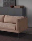 A modern beige Sonder Sofa by Wooden Stories, featuring thin black legs, is placed in front of a dark cabinet that displays a brown bowl, a rectangular box, and a framed abstract artwork against a light gray wall.