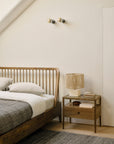 A minimalist bedroom featuring the Ethnicraft Spindle Bed in teak, white bedding, a gray blanket, a wooden nightstand with a lamp, books, decor accents, and two wall-mounted lights above the bed on a white wall.