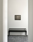A minimalist room features the Ethnicraft Spindle Bench - Black Oak set against a white wall with gray tiled floor, and a small framed abstract artwork in gray tones hung above the bench.