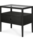 The Ethnicraft Spindle Nightstand - Black Oak features modern slim legs, a glass top, an open shelf below, and a single drawer with a round hole handle, inspired by contemporary design.