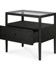 The Spindle Nightstand - Black Oak by Ethnicraft showcases a modern, minimalist design in solid black oak, featuring a glass top, open shelf, and a partially pulled-out drawer on four slender legs.