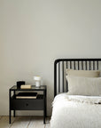 A minimal bedroom features a black metal bed frame, beige textured bedding, and an Ethnicraft Spindle Nightstand in black oak adorned with books, a small white lamp, and décor, all set against a plain light-colored wall.