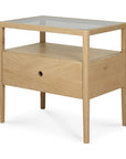 The Ethnicraft Spindle Nightstand - Oak features solid oak construction, a glass top, one open shelf, and a single drawer with a round cutout handle. Its minimalist design highlights clean lines and a light wood finish.
