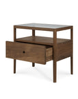 The Ethnicraft Spindle Nightstand in FSC-certified teak features slender legs, a glass top, an open shelf beneath, and a partially open drawer. The wood has a warm medium-brown finish.