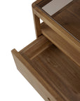 A close-up of the Ethnicraft Spindle Nightstand - Teak with a glass top shows an open drawer below. Crafted from FSC-certified teak, it features a natural finish with visible grain patterns.