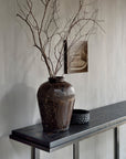 A large, textured brown vase with bare branches sits on the Ethnicraft Stability Console in Umber against a light wall, paired with a small rectangular artwork above and a black decorative bowl, evoking classic European refinement.