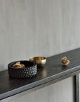 A textured black bowl, a small brass bowl, and dried seed pods from Ethnicraft grace the Stability Console - Umber, their arrangement evoking classic European refinement against a simple light gray wall.