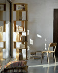 An Ethnicraft Stairs Column - Oak bookshelf, filled with books, stands against a white wall. A wooden chair faces a floor book stack nearby, while sunlight highlights a patterned cushion and the room’s elegant Ethnicraft design.