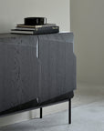 The Ethnicraft Stairs Sideboard - 3 door features European black-stained oak with angular doors and black metal legs. Topped with a stack of books and a round black dish, it sits in a light-walled room with a concrete floor.