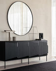 An Ethnicraft Stairs Sideboard - 4 door in black stands against a light wall, adorned with minimalist decor: a tall lamp, a geometric wire sculpture, and a small book. Above it hangs a large round mirror reflecting sheer curtains.
