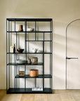 The Ethnicraft Studio Rack in black metal, with open shelves displaying ceramic bowls, jars, vases, and wooden boxes, stands against a cream wall beside a closed arched door on a wooden floor.