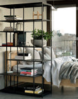 A modern bedroom features an Ethnicraft solid teak bed with gray and white bedding, an Ethnicraft Studio Rack shelving unit, books, a potted plant, and a city view through the large window in the background.
