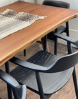 A close-up of a solid ash dining table with a woven runner and SOBU Sven Dining Chair - Black Ash on a dark wood floor, sunlight highlighting the durable lacquer finish on the tabletop and chair seat.