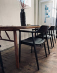A solid ash table with SOBU Sven Dining Chairs in Black Ash sits on a rustic floor. A dark vase with red branches and a small bowl rest atop, sunlight streams through tall windows, and photos are taped to a white door behind.
