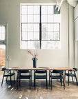 A minimalist dining room with a long solid ash wood table, six SOBU Sven Dining Chairs in black ash and leather, a black vase with tall red branches, large industrial-style windows, and sunlight streaming onto the wooden floor.