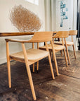 Three SOBU Sven Dining Chairs in White Ash are lined up beside a wooden table on a rustic floor. A large tumbleweed rests on the table, with photos displayed on a white door in the background.
