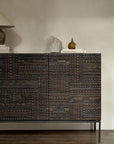 The Ethnicraft Tabwa Sideboard - 3 door, featuring dark wood and geometric motifs inspired by Tabwa culture, stands against a light beige wall, topped with stacked books, a stone sculpture, a brown vase, and a lamp with a beige shade.