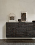 The Ethnicraft Tabwa Sideboard - 3 door, with dark, geometric carvings inspired by Tabwa culture, stands against a neutral wall and is styled with a ceramic lamp, books, pottery, and framed abstract art for a modern earthy look.