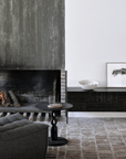 A modern living room with a lit fireplace, gray armchair, and round black side table features the Ethnicraft Tabwa TV Cupboard – 94 as a low black console displaying art, a bowl, and a lamp. Exposed brick flooring complements the minimalist look.