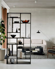A modern living room features a black open Ethnicraft shelving unit displaying decor and dividing the space. In the background, a beige sofa, rug, floor lamp, indoor plants by a large window, and Ethnicrafts Teak Abstract Coffee Table are visible.