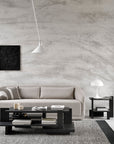 Modern minimalist living room with a beige sofa, Ethnicraft Teak Abstract Side Table, side table with a white lamp, abstract wall art, and large hanging lamp—radiating European refinement against a textured gray wall and light rug.