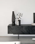 An Ethnicraft Graphic TV Cupboard - 63 with geometric patterns holds tall dark vases and a smaller light vase with dried branches, positioned against a plain white wall in a minimalist room.
