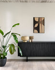 A modern living space features a large potted plant, the Ethnicraft Mosaic Sideboard - 3 doors displaying fine craftsmanship, neutral ceramic vases, and an abstract geometric art piece on a white wall.