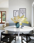 A sunlit dining area features a white oval table, two SOBU Thor Dining Chairs in black ash with woven paper cord seats, a blue vase with yellow-green foliage, a small bowl, a gray sofa, and abstract black-and-white artwork.